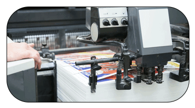 A person operates a large-format printer, feeding paper through the machine while multiple ink cartridges are visible in the foreground. The printer appears to be processing a print job on white paper.