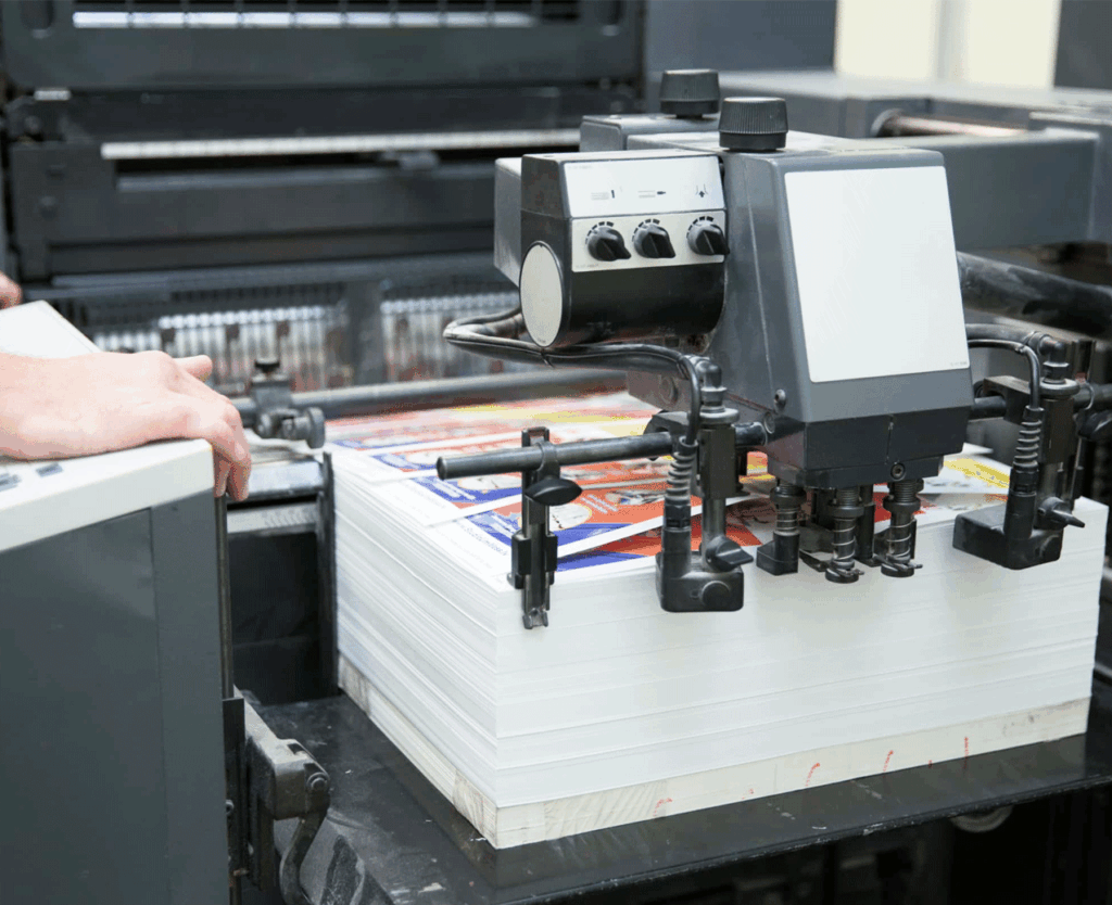A person operates a large-format printer, feeding paper through the machine while multiple ink cartridges are visible in the foreground. The printer appears to be processing a print job on white paper.